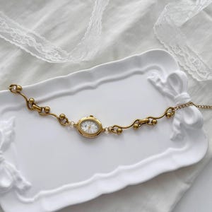 May include: A gold-coloured bracelet watch with an oval face and a white dial. The watch is set on a decorative white tray. The bracelet features a delicate, flowing design with small, round accents. The watch is a vintage-inspired accessory.
