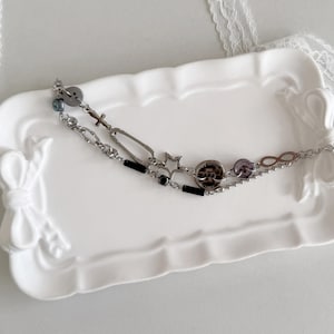 Pode incluir: Uma pulseira prateada em camadas com vários berloques, incluindo uma cruz, uma estrela e um símbolo de infinito. A pulseira também apresenta contas pretas e roxas, e é exibida em uma bandeja decorativa branca com detalhes de laço.