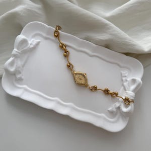 May include: A gold-toned vintage wristwatch with an oval face and delicate chain. The watch rests on a white, bow-shaped tray. The watch band features small, spherical accents. The background is a white, textured fabric.