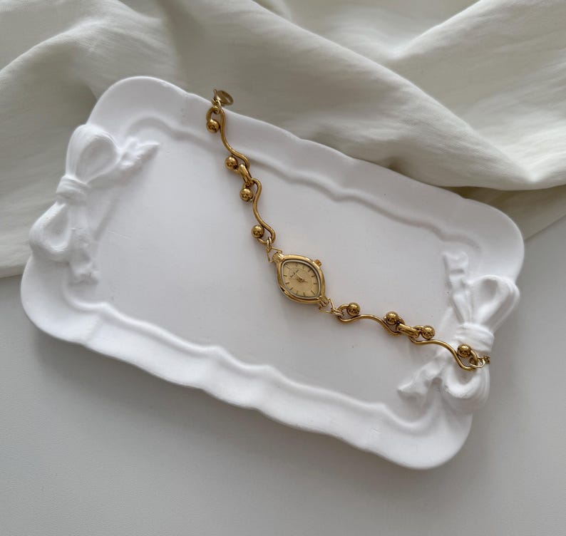 May include: A gold-toned vintage wristwatch with an oval face and delicate chain. The watch rests on a white, bow-shaped tray. The watch band features small, spherical accents. The background is a white, textured fabric.