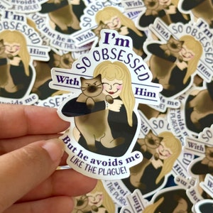 May include: A sticker with a cartoon illustration of a woman with blonde hair holding a cat. The text on the sticker reads "I'm so obsessed with him, but he avoids me like the plague!"