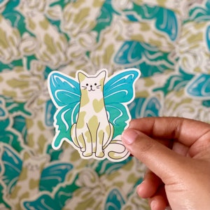 May include: A white cat with yellow spots wearing blue butterfly wings. The cat is sitting with its tail curled around its legs. The wings are a gradient of blue and green.