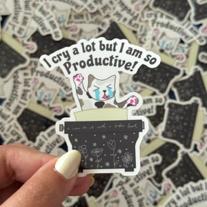 May include: A white sticker with a black and white cat crying and typing on a typewriter. The sticker says "I cry a lot but I am so productive!" and "I can do it well with a broken heart."