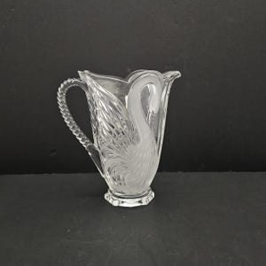 May include: A clear glass jug featuring a frosted swan design on the side. The jug has a textured, rope-like handle and a scalloped rim. The body of the swan is detailed with etched feathers, and the base of the jug is faceted.