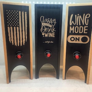 Op de afbeelding: Drie houten wijnboxdispensers met zwarte voorkanten en natuurlijke houten frames. Eén heeft een Amerikaans vlagontwerp, een andere zegt "Classy Bitches Drink Wine", en de derde zegt "Wine Mode On".