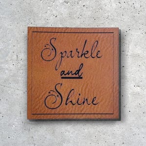 Könnte beinhalten: Ein braunes Lederstück mit dem Text "Sparkle and Shine" in schwarzer Schrift.
