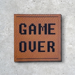 Könnte beinhalten: Ein braunes Leder-Patch mit schwarzem, pixeligem Text, der "GAME OVER" lautet.