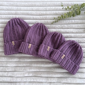 May include: Four purple crochet beanies arranged in a row, each featuring a ribbed texture and a small, rectangular tan leather-like tag attached to the folded brim. The hats are identical in colour and design, showcasing a consistent knit pattern.