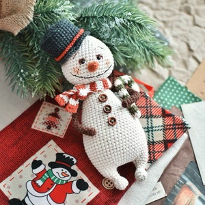 Puede incluir: Un mu&ntilde;eco de nieve de crochet con un sombrero negro, una bufanda marr&oacute;n y un bast&oacute;n de caramelo en la mano. El mu&ntilde;eco de nieve est&aacute; sentado sobre una tela de cuadros rojos y blancos con una tela de lunares verdes detr&aacute;s.