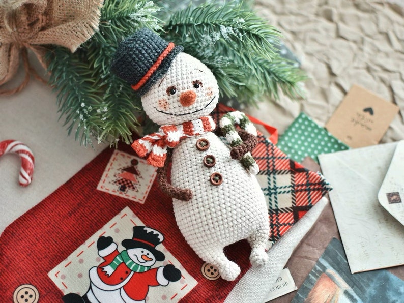 Puede incluir: Un mu&ntilde;eco de nieve de crochet con un sombrero negro, una bufanda marr&oacute;n y un bast&oacute;n de caramelo en la mano. El mu&ntilde;eco de nieve est&aacute; sentado sobre una tela de cuadros rojos y blancos con una tela de lunares verdes detr&aacute;s.