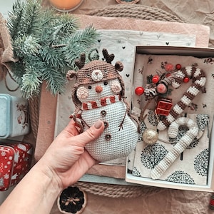 May include: A crocheted snowman ornament with a brown reindeer hat and a red and white scarf. The ornament is in a hand, and a box with a crocheted candy cane and other Christmas decorations is in the background.