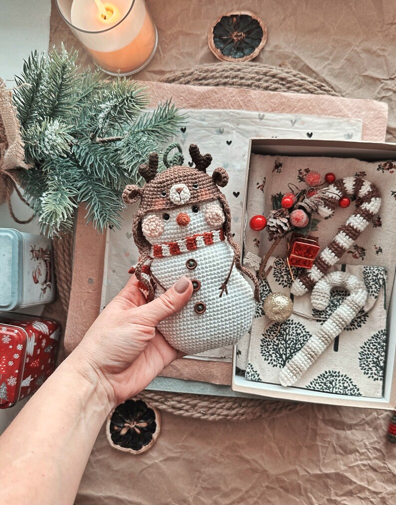 May include: A crocheted snowman ornament with a brown reindeer hat and a red and white scarf. The ornament is in a hand, and a box with a crocheted candy cane and other Christmas decorations is in the background.