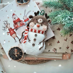 May include: A crocheted snowman ornament with a brown hat, red and white striped scarf, and brown antlers. The snowman is sitting on a white and brown fabric background.