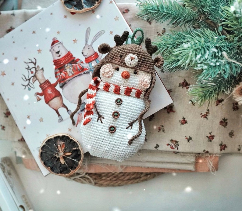 May include: A crocheted snowman ornament with a brown hat, red and white striped scarf, and brown antlers. The snowman is sitting on a white and brown fabric background.