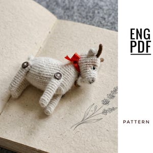 May include: A small, off-white crocheted goat figurine with brown button joints at the legs, a red ribbon tied around its neck, and small brown horns. The text ENG PDF PATTERN is visible on the right side of the image.