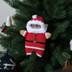 May include: A crocheted Santa Claus ornament with a white beard, red and white striped suit, and black belt. The ornament is hanging on a green Christmas tree.