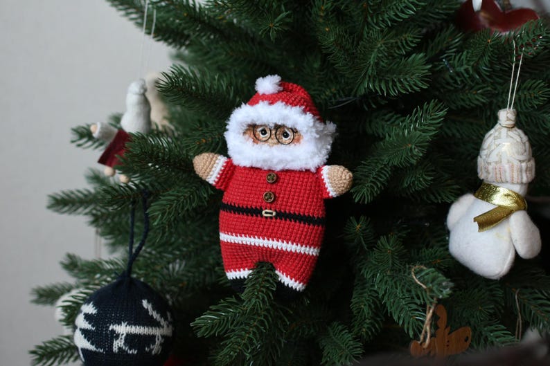 May include: A crocheted Santa Claus ornament with a white beard, red and white striped suit, and black belt. The ornament is hanging on a green Christmas tree.