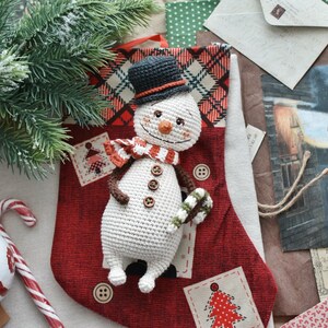Puede incluir: Un adorno de mu&ntilde;eco de nieve de crochet con un sombrero de copa negro, una bufanda roja y blanca y un bast&oacute;n de caramelo en la mano. El mu&ntilde;eco de nieve est&aacute; sentado en una media roja y blanca con un patr&oacute;n de cuadros.