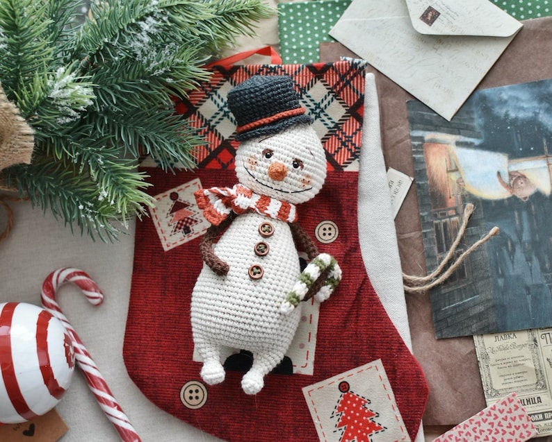 Puede incluir: Un adorno de mu&ntilde;eco de nieve de crochet con un sombrero de copa negro, una bufanda roja y blanca y un bast&oacute;n de caramelo en la mano. El mu&ntilde;eco de nieve est&aacute; sentado en una media roja y blanca con un patr&oacute;n de cuadros.