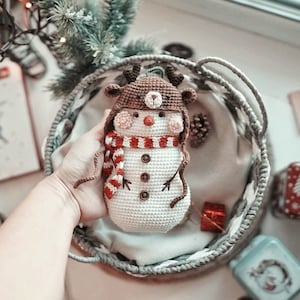 May include: A crocheted snowman ornament with a brown reindeer hat, red and white striped scarf, and brown button eyes. The snowman is sitting in a gray wicker basket.