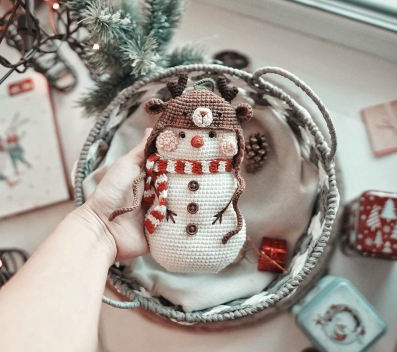 May include: A crocheted snowman ornament with a brown reindeer hat, red and white striped scarf, and brown button eyes. The snowman is sitting in a gray wicker basket.