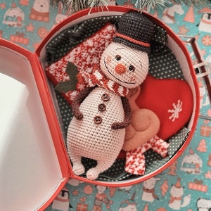Puede incluir: Un juguete de mu&ntilde;eco de nieve de crochet con un sombrero marr&oacute;n y una bufanda roja, sentado dentro de una caja de regalo roja con un forro estampado blanco y rojo. La caja est&aacute; sobre una superficie estampada azul y blanca.
