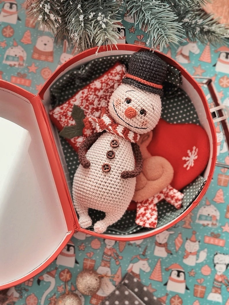 Puede incluir: Un juguete de mu&ntilde;eco de nieve de crochet con un sombrero marr&oacute;n y una bufanda roja, sentado dentro de una caja de regalo roja con un forro estampado blanco y rojo. La caja est&aacute; sobre una superficie estampada azul y blanca.