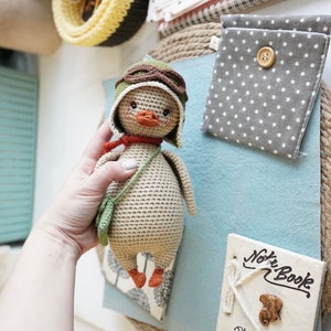 Pode incluir: Um pato de croch&ecirc; usando um chap&eacute;u de aviador marrom com &oacute;culos, uma echarpe verde e uma bolsa de croch&ecirc; verde. O pato &eacute; bege com p&eacute;s laranja.