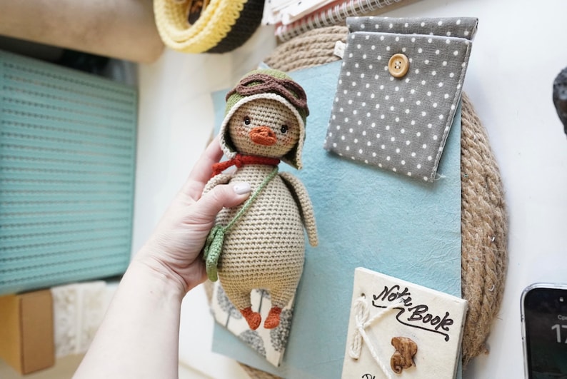 Pode incluir: Um pato de croch&ecirc; usando um chap&eacute;u de aviador marrom com &oacute;culos, uma echarpe verde e uma bolsa de croch&ecirc; verde. O pato &eacute; bege com p&eacute;s laranja.