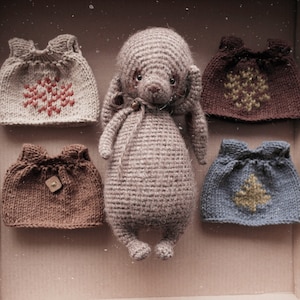 May include: A grey crocheted bunny toy with long ears and a brown nose, surrounded by five knitted doll clothes in various colours and patterns. The clothes have different designs, including snowflakes, trees, and geometric patterns.