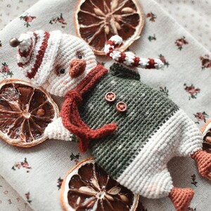 May include: A crocheted snowman toy with a red and white hat, green and white body, and a red scarf. The toy is sitting on a white fabric background with dried orange slices.