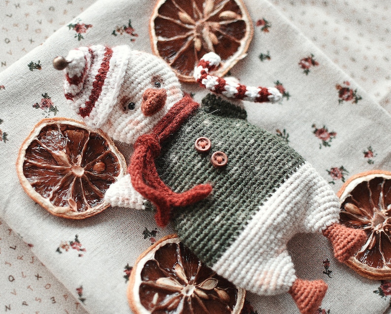 May include: A crocheted snowman toy with a red and white hat, green and white body, and a red scarf. The toy is sitting on a white fabric background with dried orange slices.