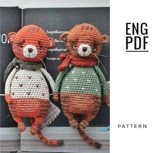 Könnte beinhalten: Zwei gehäkelte Tiger-Amigurumi-Spielzeuge, die Pullover und Schals tragen. Der Tiger links trägt einen beigefarbenen Pullover mit roten Herzen und einen grünen Schal. Der Tiger rechts trägt einen grünen Pullover mit weißen Herzen und einen roten Schal. Beide Tiger haben orange und braune Streifen.