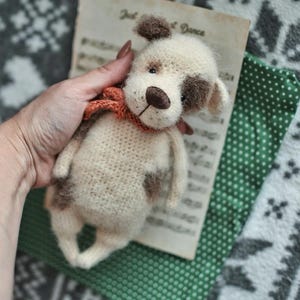 May include: A crocheted stuffed dog toy with brown and white fur, wearing a red and brown scarf. The dog is being held in a hand.