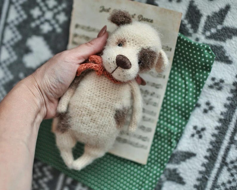 May include: A crocheted stuffed dog toy with brown and white fur, wearing a red and brown scarf. The dog is being held in a hand.