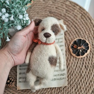 May include: A crocheted stuffed dog toy with white and brown fur, wearing a red and orange collar. The toy is sitting on a sheet of music.