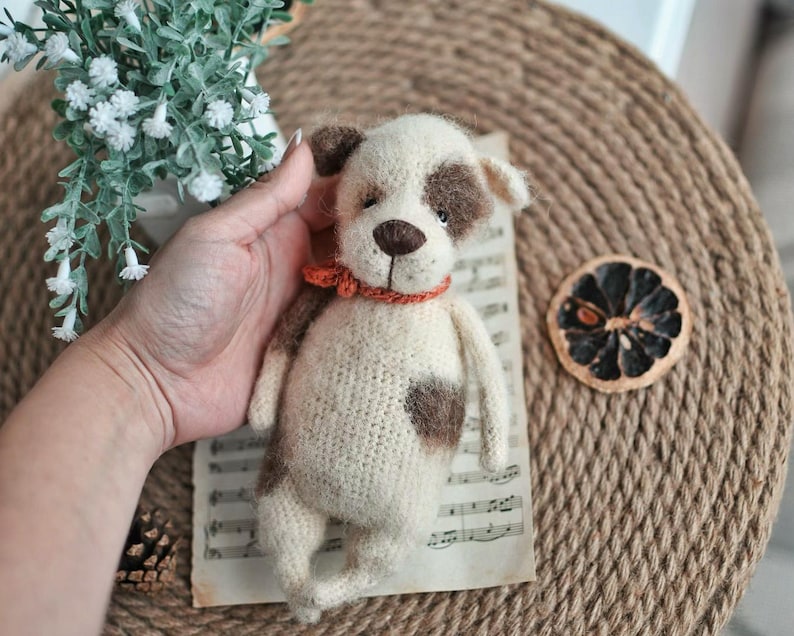 May include: A crocheted stuffed dog toy with white and brown fur, wearing a red and orange collar. The toy is sitting on a sheet of music.