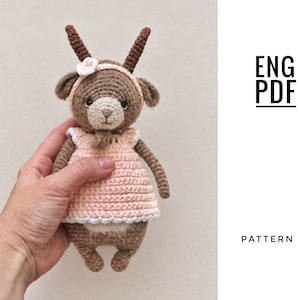 May include: A brown crocheted goat doll with a light beige muzzle, wearing a pale pink dress with a white scalloped trim and a matching headband with a small white flower. The text ENG PDF PATTERN is visible on the right side of the image.