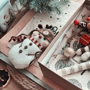 May include: A crocheted snowman with a red and white striped scarf and a brown crocheted reindeer hat is lying in a brown gift box. The box is open and contains a crocheted reindeer ornament and a small red gift box.