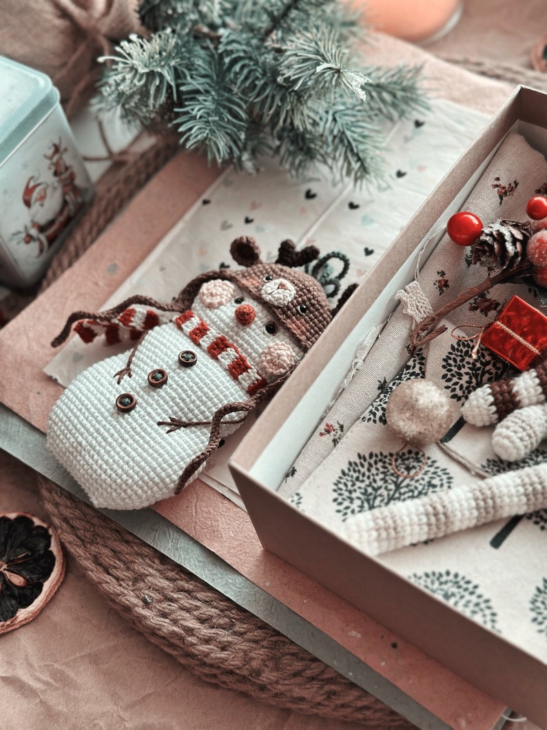 May include: A crocheted snowman with a red and white striped scarf and a brown crocheted reindeer hat is lying in a brown gift box. The box is open and contains a crocheted reindeer ornament and a small red gift box.