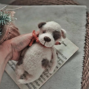 May include: A crocheted stuffed animal dog with white and brown fur, wearing a red and orange bow tie. The dog is sitting on a piece of sheet music.