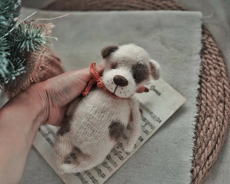 May include: A crocheted stuffed animal dog with white and brown fur, wearing a red and orange bow tie. The dog is sitting on a piece of sheet music.