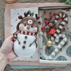 May include: A crocheted snowman wearing a reindeer hat with brown antlers and a red and white striped scarf. The snowman is sitting in a gift box with a crocheted candy cane and other Christmas decorations.