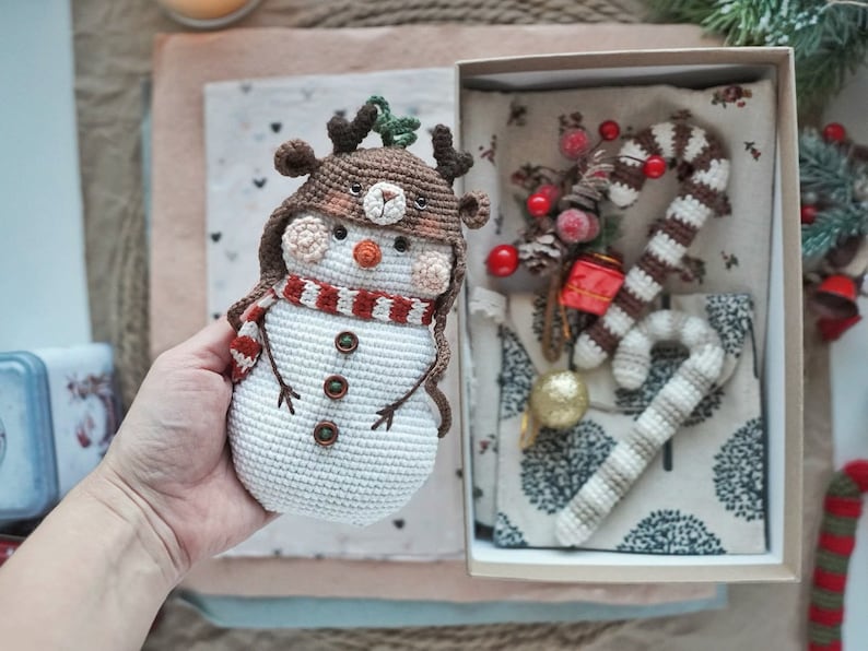 May include: A crocheted snowman wearing a reindeer hat with brown antlers and a red and white striped scarf. The snowman is sitting in a gift box with a crocheted candy cane and other Christmas decorations.