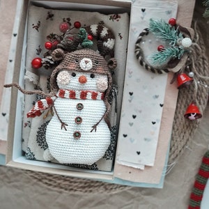 May include: A crocheted snowman with a brown and white hat, red and white scarf, and brown button eyes. The snowman is in a box with a white and gray patterned fabric background.