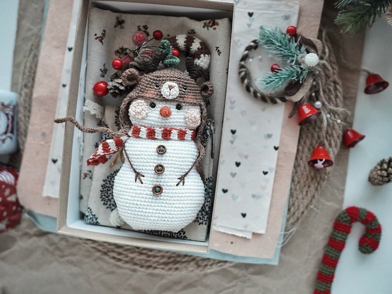 May include: A crocheted snowman with a brown and white hat, red and white scarf, and brown button eyes. The snowman is in a box with a white and gray patterned fabric background.