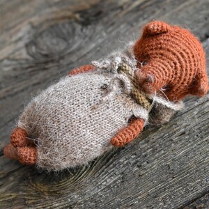 Op de afbeelding: Een handgemaakte, gebreide pop in liggende positie. De pop heeft een bruin hoofd en ledematen, met een lichtbruin lichaam en een kleine, gedetailleerde outfit. De pop ligt op een verweerd houten oppervlak.
