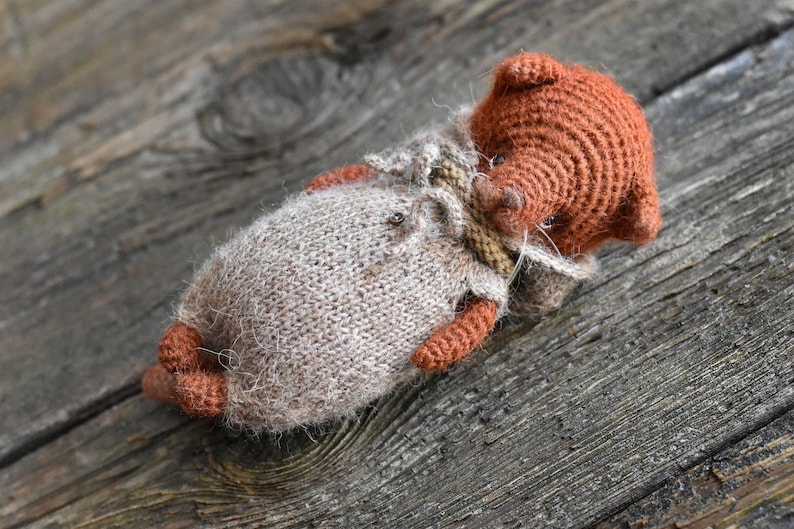 Op de afbeelding: Een handgemaakte, gebreide pop in liggende positie. De pop heeft een bruin hoofd en ledematen, met een lichtbruin lichaam en een kleine, gedetailleerde outfit. De pop ligt op een verweerd houten oppervlak.