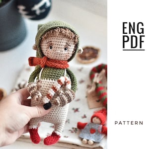 May include: Crocheted elf doll with a green hat, red scarf, and brown hair. The elf is holding a candy cane in its hand. The doll is standing on a white surface with a red and white striped candy cane in the background. ENG PDF PATTERN.