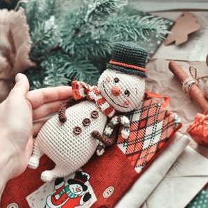 Puede incluir: Un adorno de mu&ntilde;eco de nieve de crochet con un sombrero negro, un cuerpo blanco y una bufanda a rayas rojas y blancas. El mu&ntilde;eco de nieve est&aacute; tumbado sobre una tela de cuadros rojos y blancos con un adorno de mu&ntilde;eco de nieve m&aacute;s peque&ntilde;o sobre la tela.